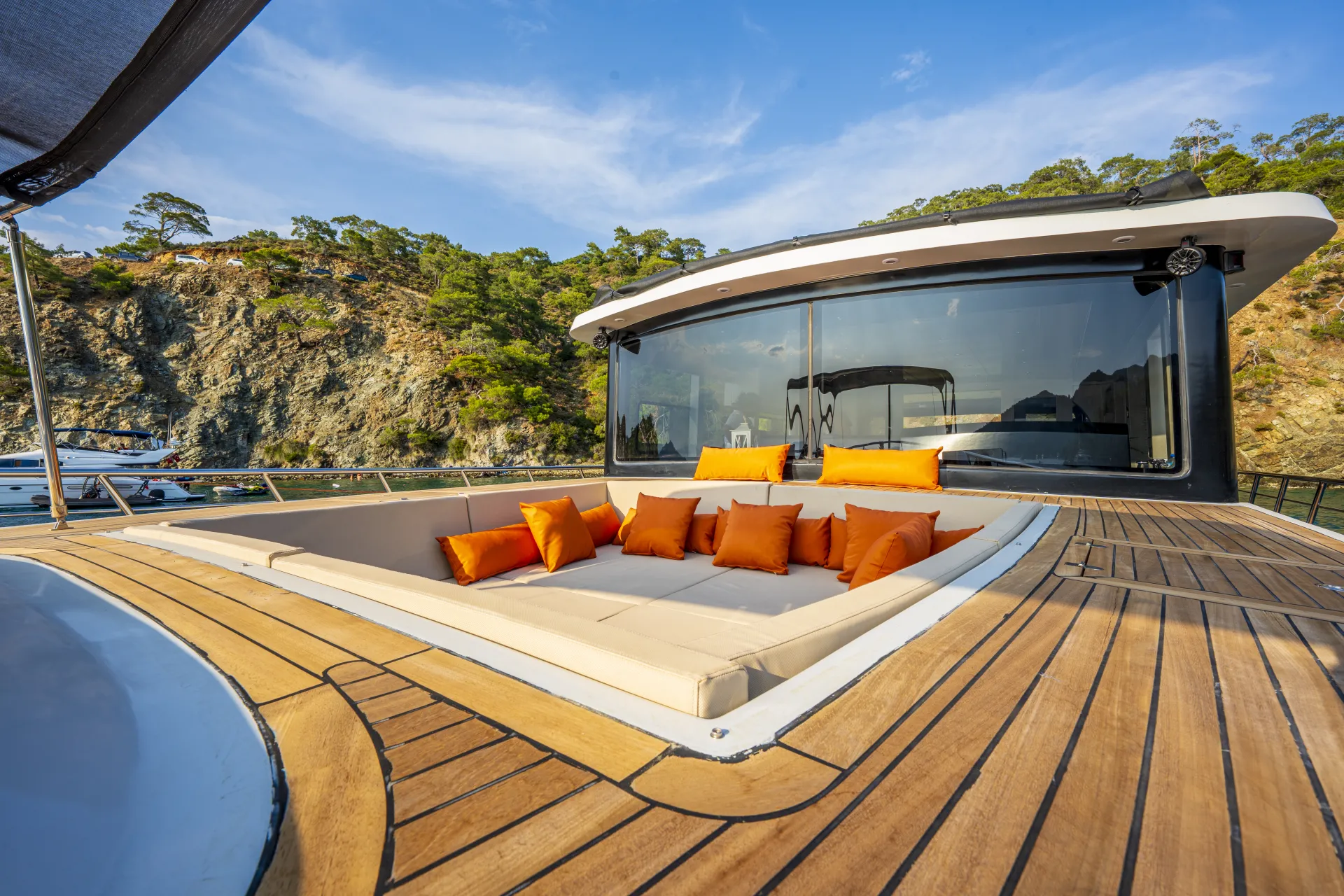 JUNIOR SIMAY A luxurious yacht deck features a sunken lounge area with beige cushions and bright orange pillows, set against a backdrop of rocky cliffs, green trees, and calm blue water.