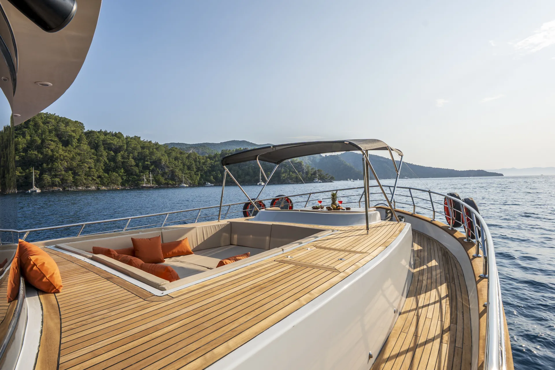 JUNIOR SIMAY A luxury yacht deck with wooden flooring, orange cushions on a built-in seating area, and a canopy, sailing near a forested coastline under a clear sky.
