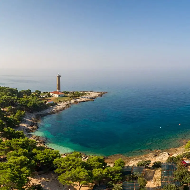 A lighthouse stands on a rocky peninsula surrounded by turquoise sea and dense green trees under a clear blue sky.