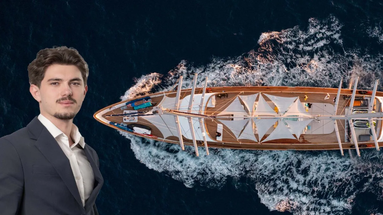 A man in a suit stands in front of an aerial view of a sailing yacht moving through dark blue ocean water, creating white waves.