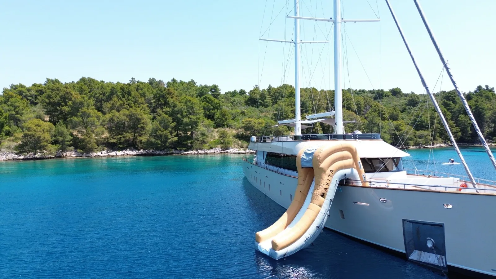 MAXITA A large yacht anchored near a forested shoreline features a beige inflatable slide extending from its upper deck into the clear, blue water. The scene is bright and peaceful under a clear sky.