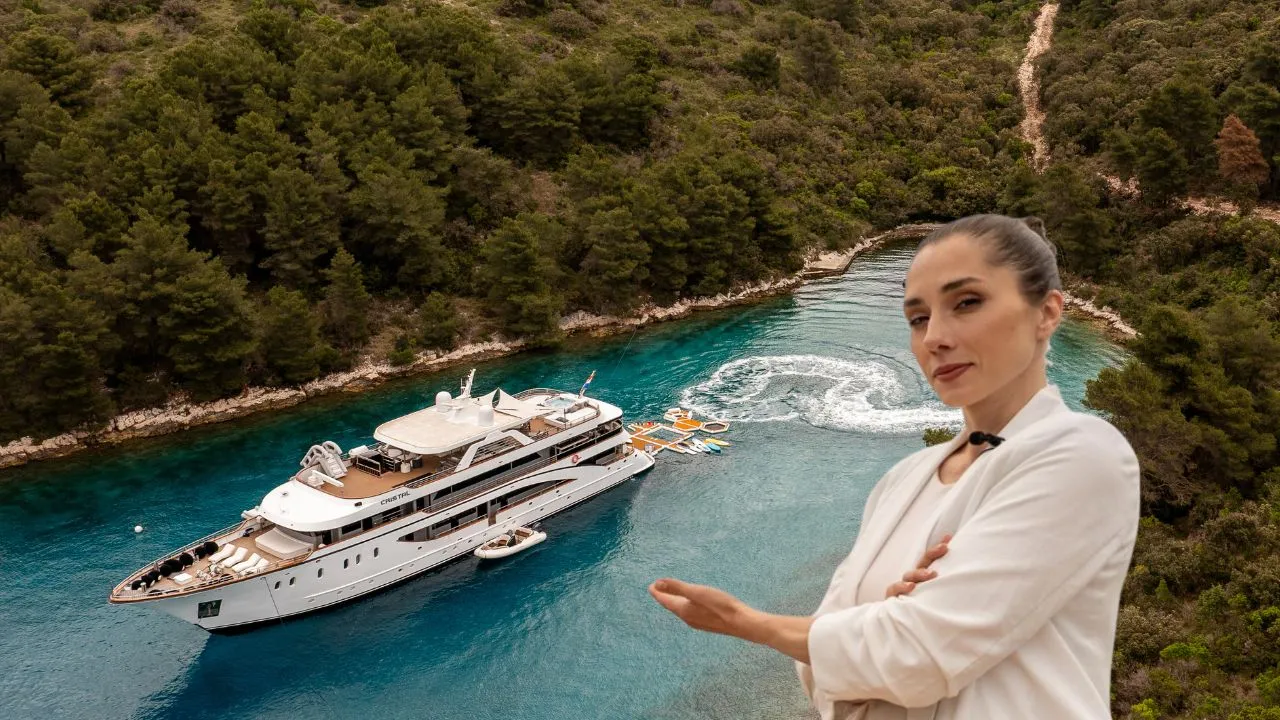 A woman in a white blazer gestures toward a large luxury yacht anchored in clear blue water near a forested coastline. The scene is viewed from above, showing the yacht, small boats, and lush greenery.