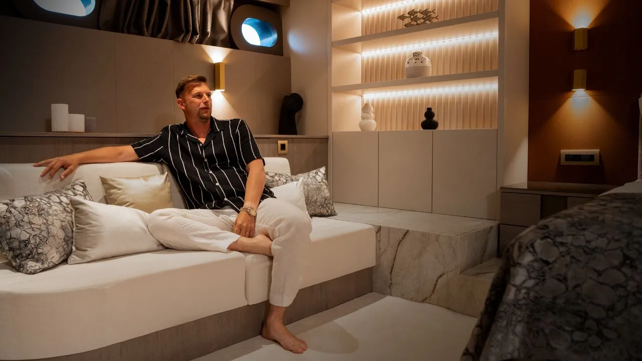 A man in a striped shirt and white pants sits barefoot on a modern white couch in a stylish, warmly lit room with built-in shelves, decorative items, and soft accent lighting.