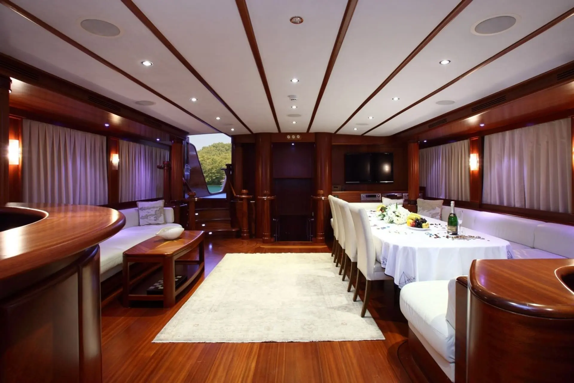 Luxurious yacht interior with polished wood floors, white couches, a dining table set with flowers and wine, soft lighting, a TV on the wall, and large windows with curtains. The space looks elegant and spacious.