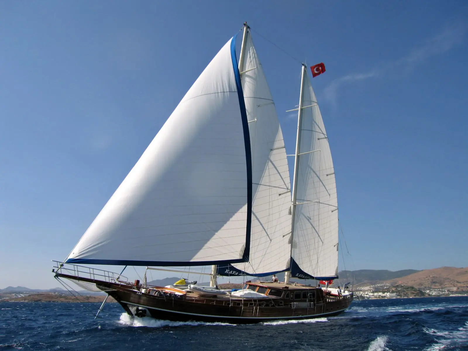 A large sailboat with two white sails moves quickly across deep blue water under a clear sky, with a red flag flying and distant hills in the background.