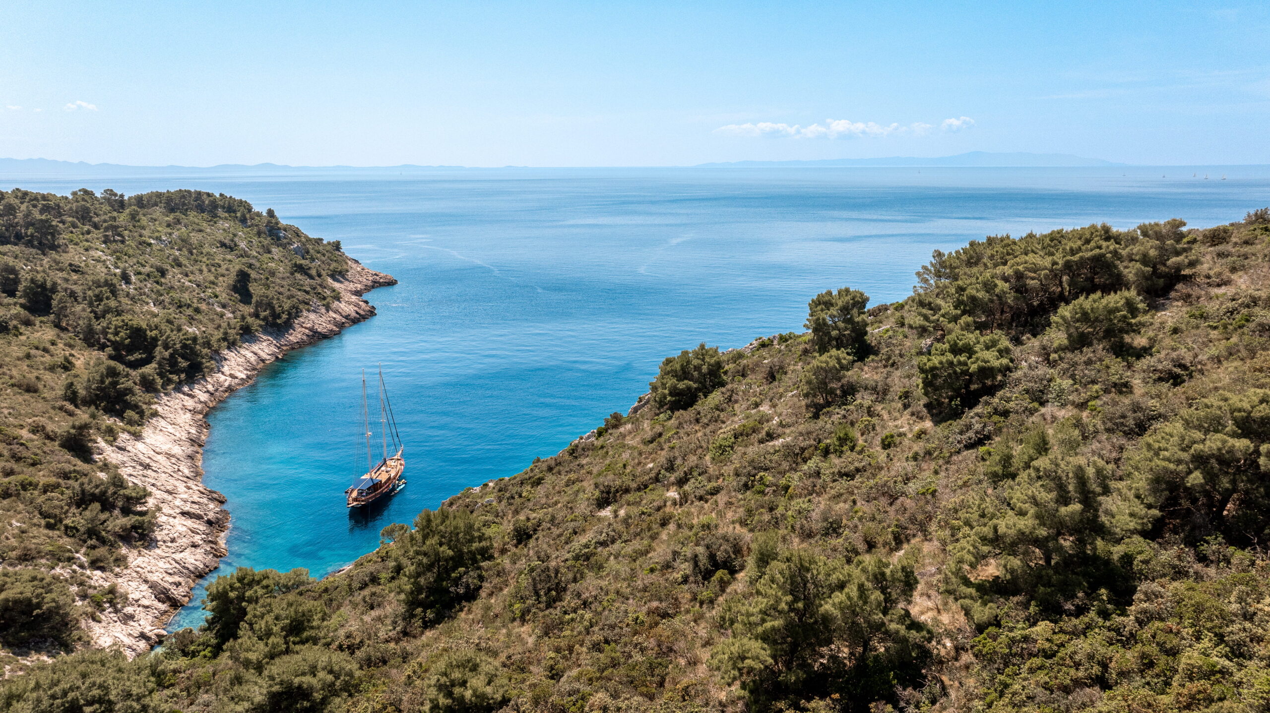 SMART SPIRIT Aerial view of a sailboat anchored in a small, secluded cove with clear blue water, surrounded by green, rocky hills under a bright, clear sky.