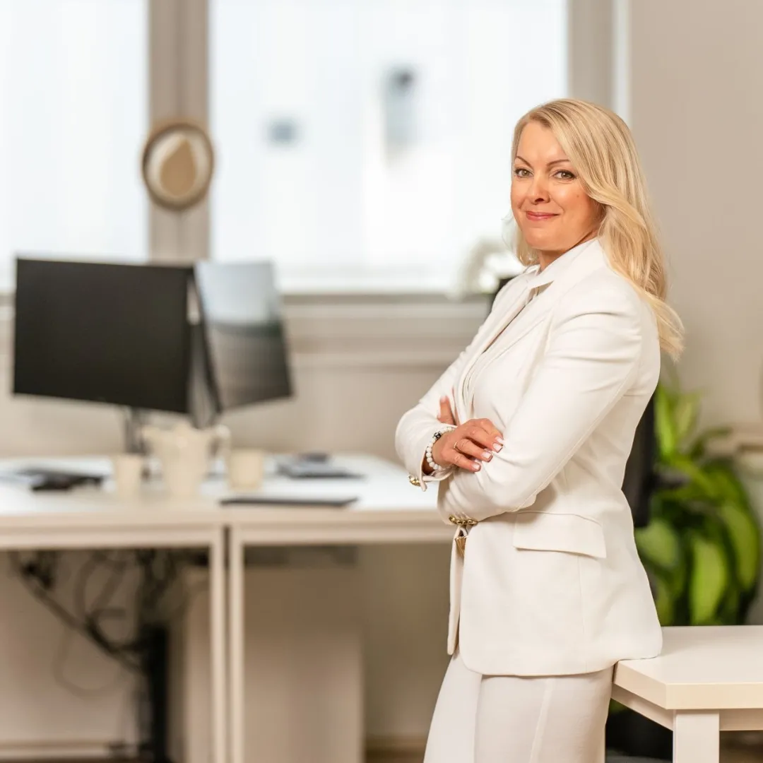 A confident woman in a white suit stands with arms crossed in a bright modern office, smiling at the camera. There are computer monitors, plants, and desks in the background.