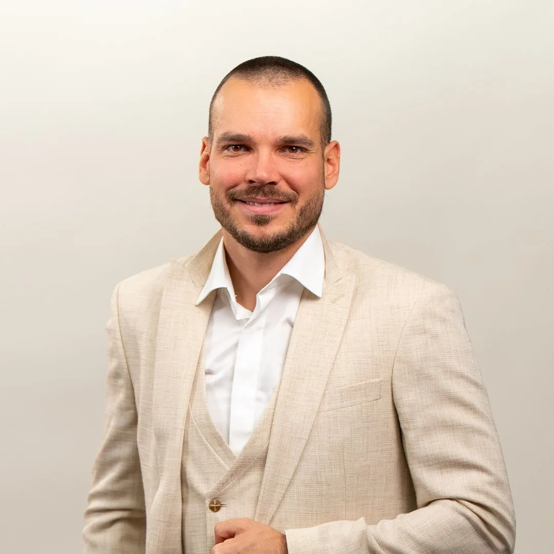 A man with a shaved head and trimmed beard smiles at the camera. He is wearing a light beige suit with a white dress shirt, standing against a plain, light background.