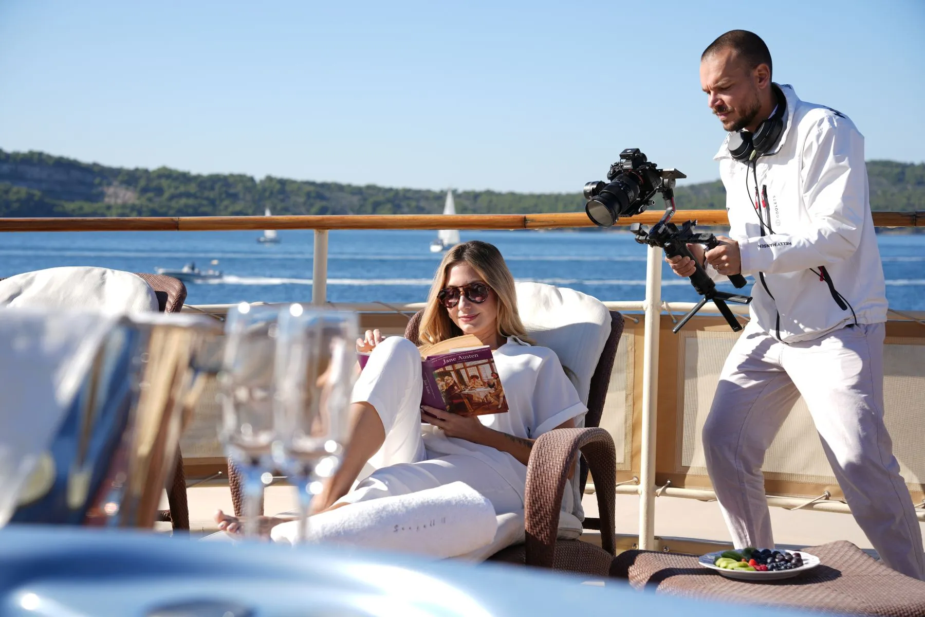 A woman relaxes on a lounge chair reading a book on a yacht, while a cameraman films her. The yacht is on calm water with a scenic view of trees and sailboats in the background.
