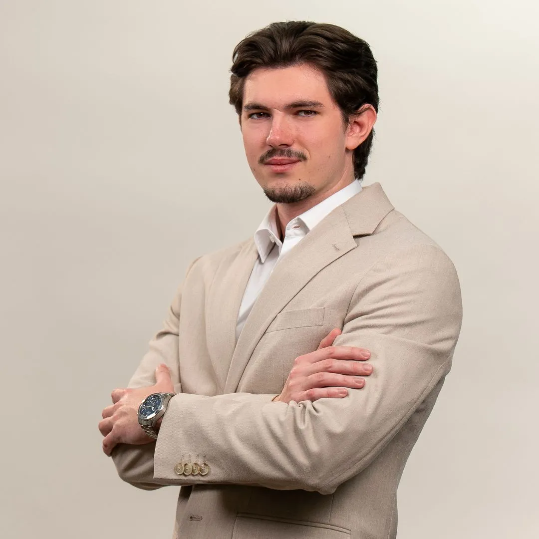 A man with dark hair and a short beard, dressed in a beige suit and white shirt, stands with arms crossed and a confident expression against a plain light background.