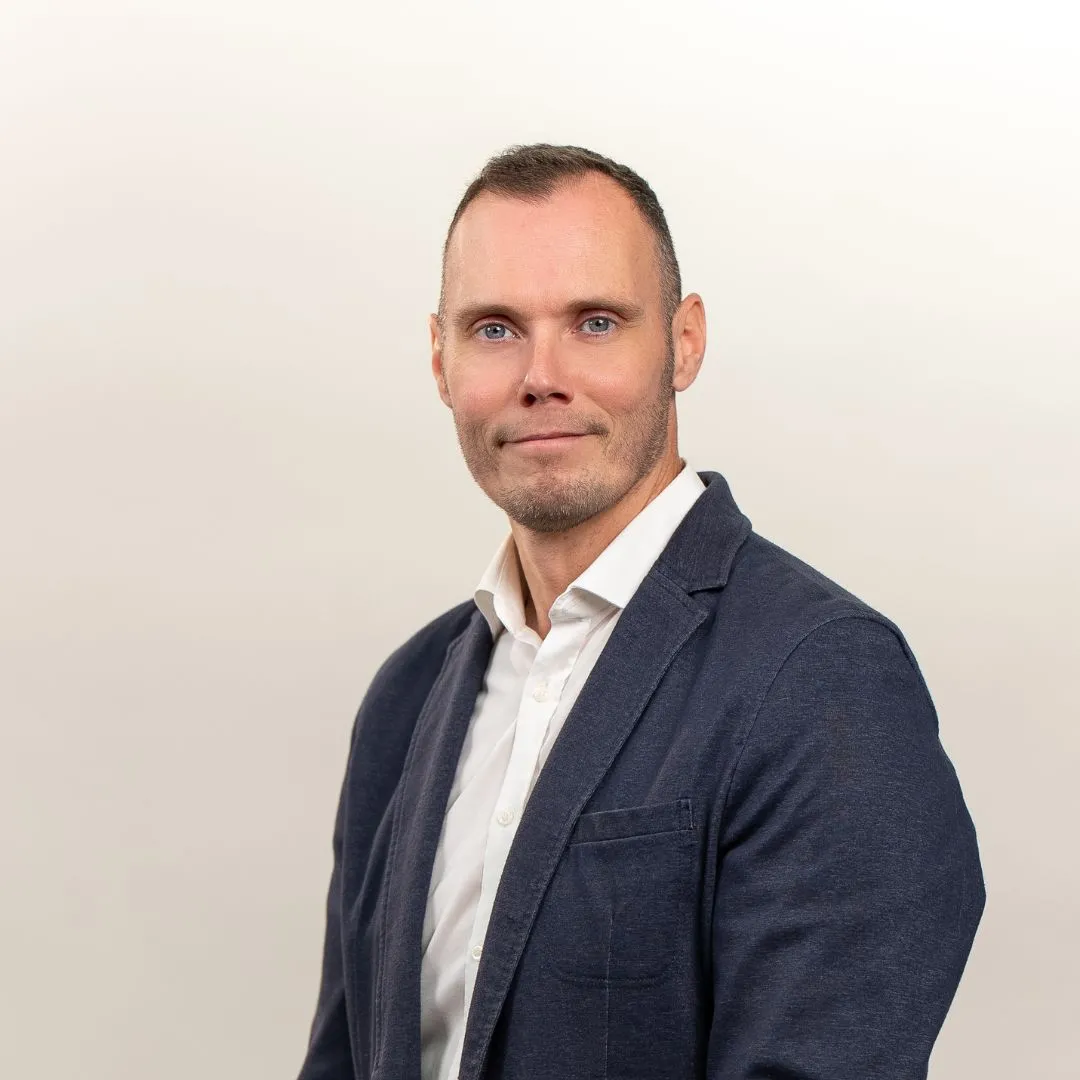 A man with short hair, wearing a white button-up shirt and a dark blazer, poses in front of a plain, light-colored background. He is looking at the camera and smiling slightly.