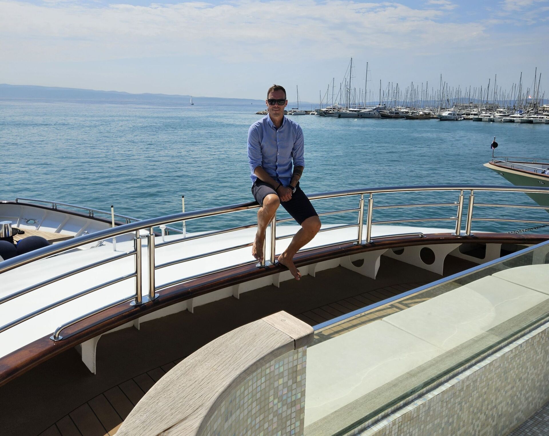 A man in a blue shirt and dark shorts sits barefoot on the railing of a yacht, with a marina full of sailboats and the blue sea visible in the background under a partly cloudy sky.