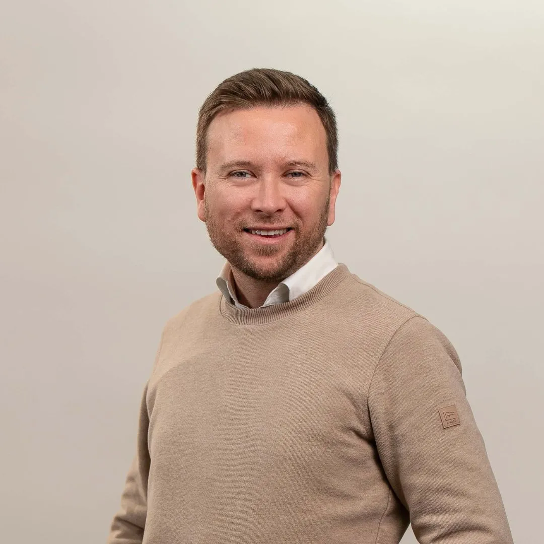 A man with short light brown hair and a beard smiles at the camera. He is wearing a beige sweater over a white collared shirt, standing against a plain light-colored background.