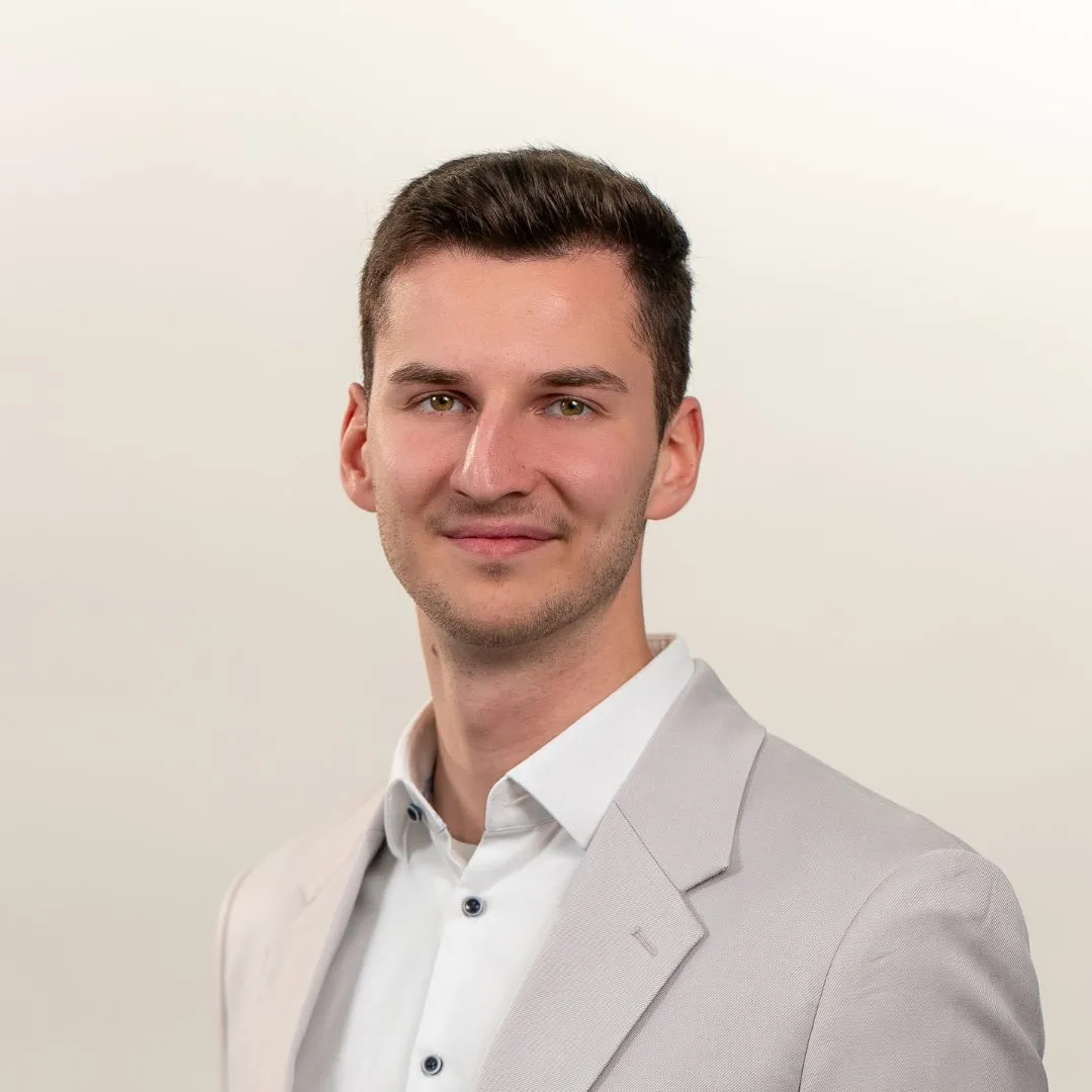 A young man with short brown hair, wearing a light grey suit jacket and white shirt, smiles slightly at the camera against a plain, light background.