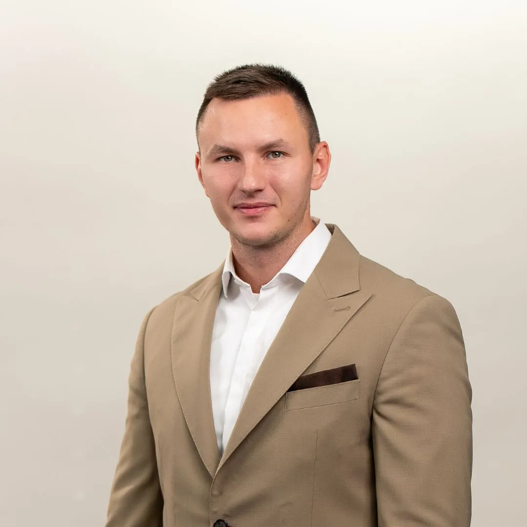A man in a tan suit jacket and white shirt stands against a plain, light background, looking at the camera with a slight smile.