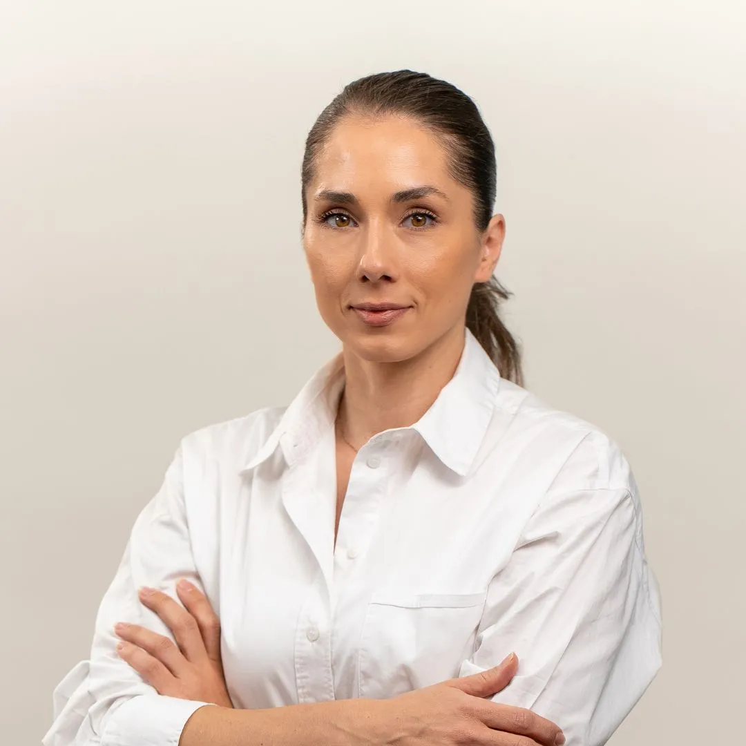 A woman with long brown hair tied back, wearing a white button-up shirt, stands with arms crossed and looks confidently at the camera against a plain light background.