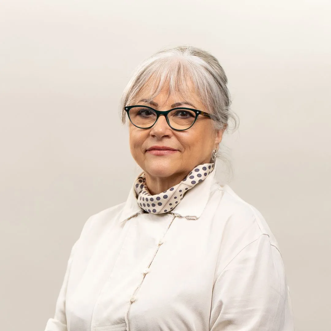 An older woman with gray hair, glasses, and a light polka dot scarf, wearing a white buttoned shirt, stands against a plain light background, looking at the camera with a slight smile.