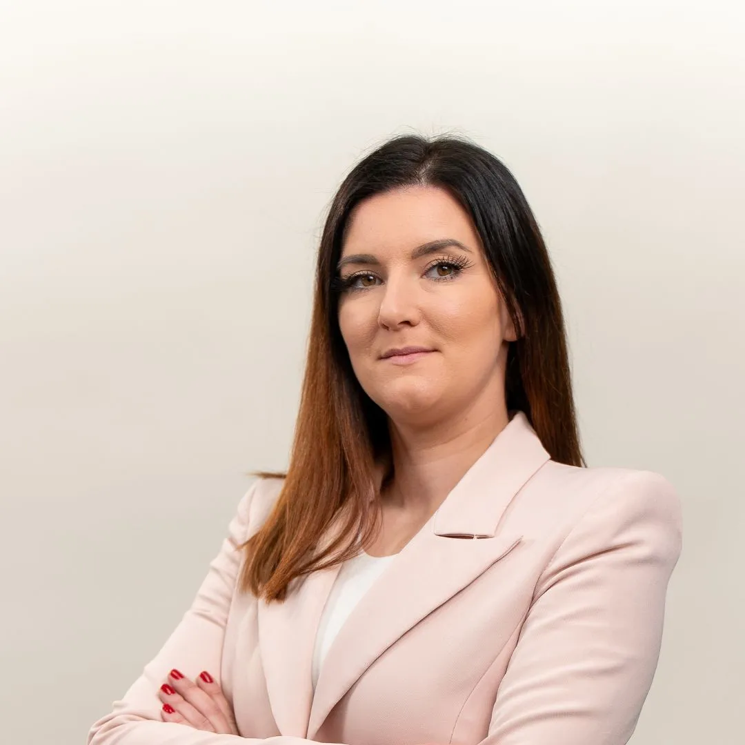 A woman with straight brown hair, dressed in a pale pink blazer and white top, stands with arms crossed and a confident expression against a plain light background.