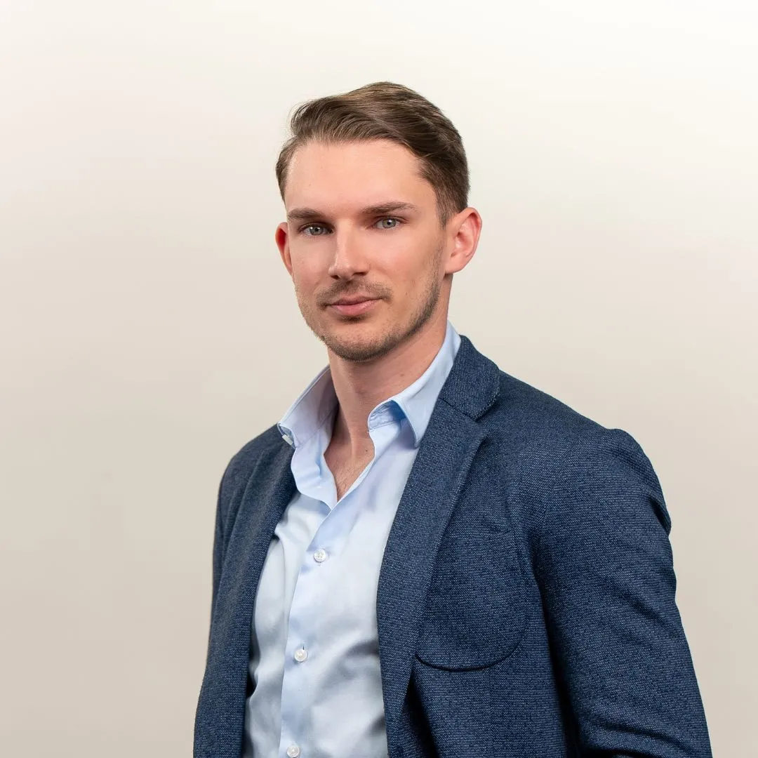 A young man with short brown hair wearing a blue blazer over a light blue button-up shirt stands against a plain, light background, looking confidently at the camera.