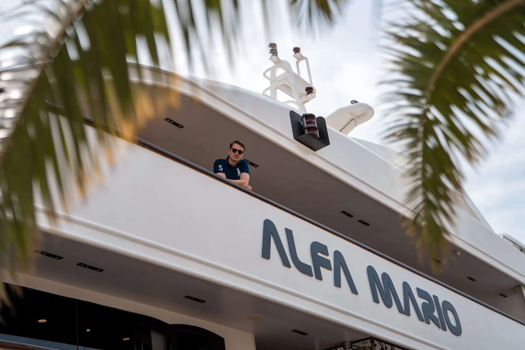 A man wearing sunglasses stands on the deck of a large white yacht named “ALFA MARIO,” partially framed by palm leaves in the foreground.