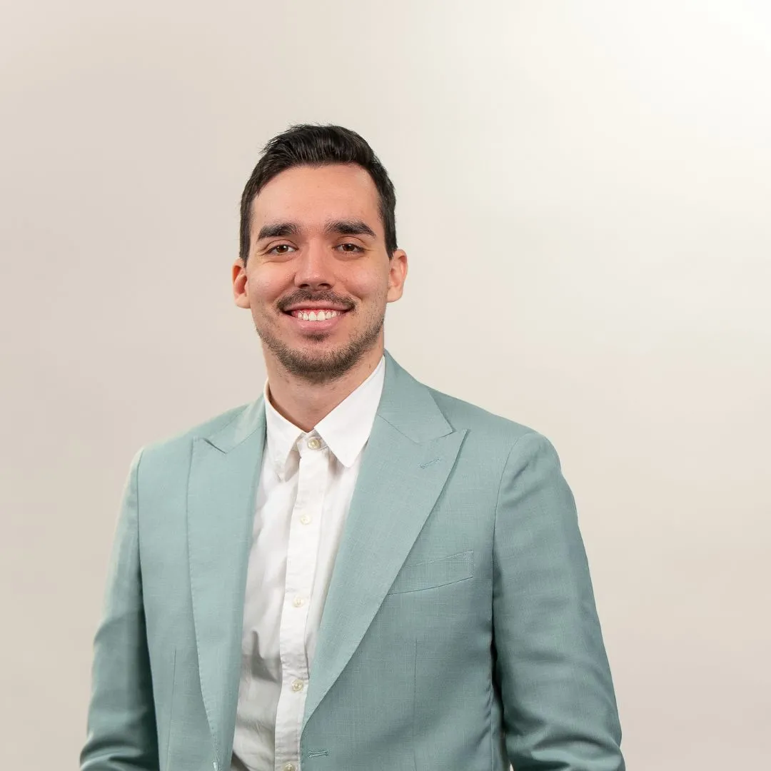 A man with short dark hair and a trimmed beard is smiling, wearing a light blue suit jacket and a white shirt, standing against a plain light background.