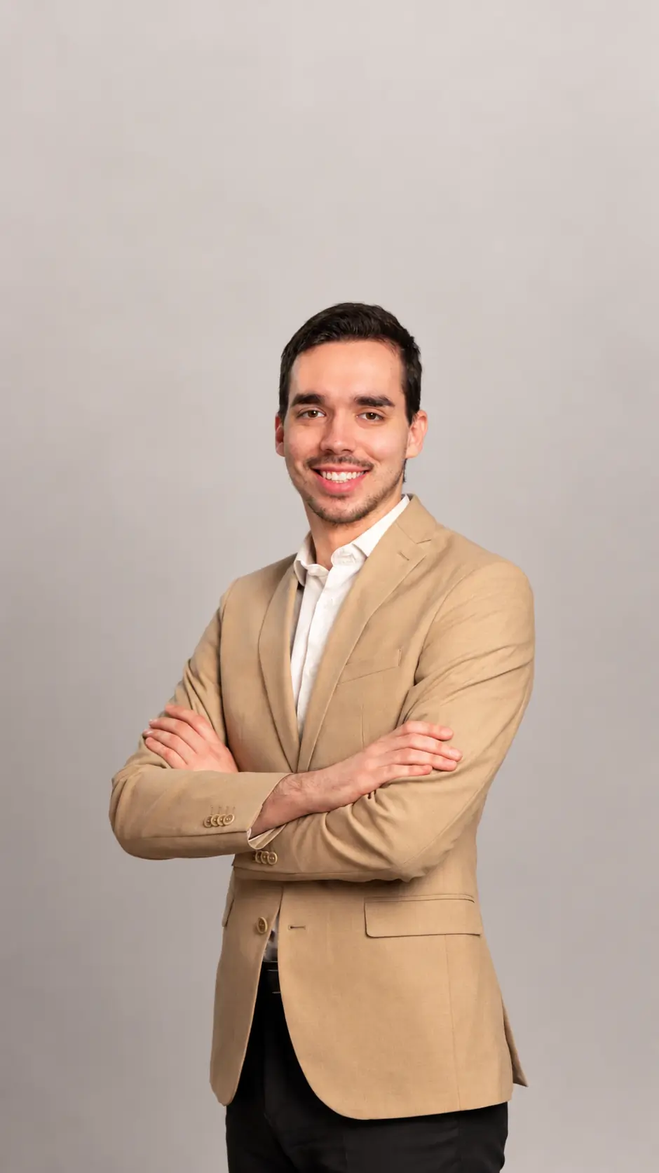 A man in a beige blazer and white shirt stands with arms crossed, smiling confidently against a plain light gray background.