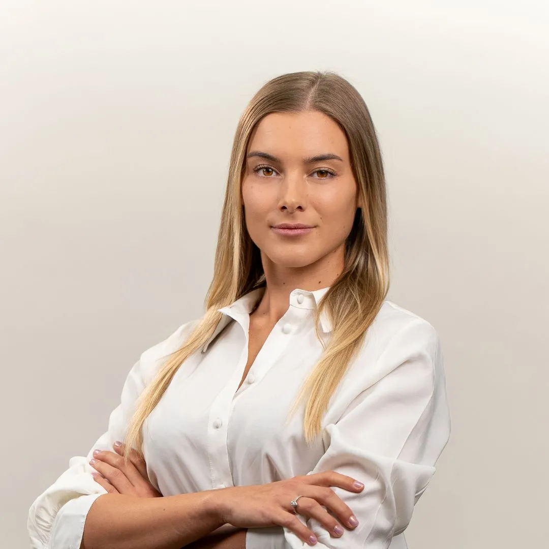 A woman with long blonde hair wearing a white button-up shirt stands with arms crossed, looking confidently at the camera against a plain light background.