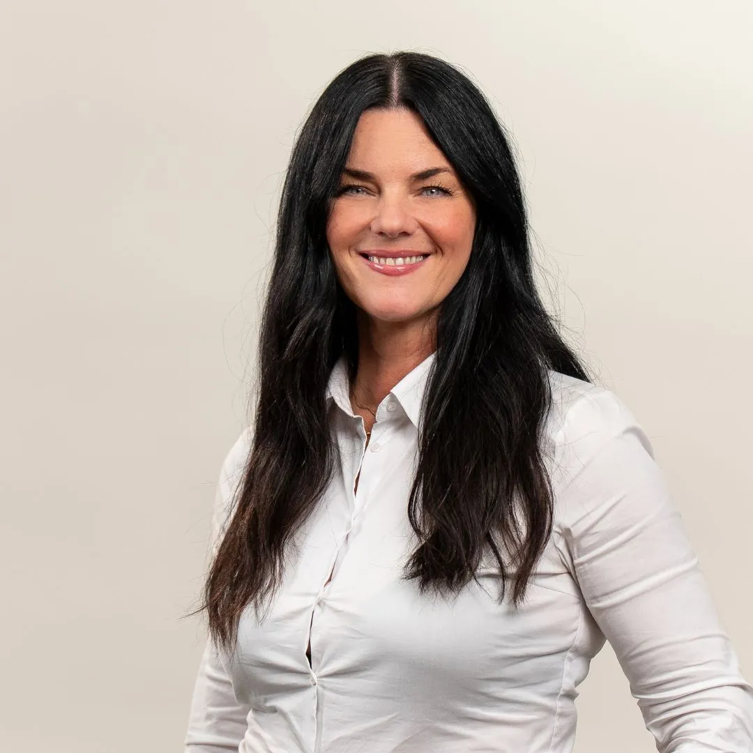 A woman with long dark hair, wearing a white button-up shirt, smiles at the camera against a plain light background.