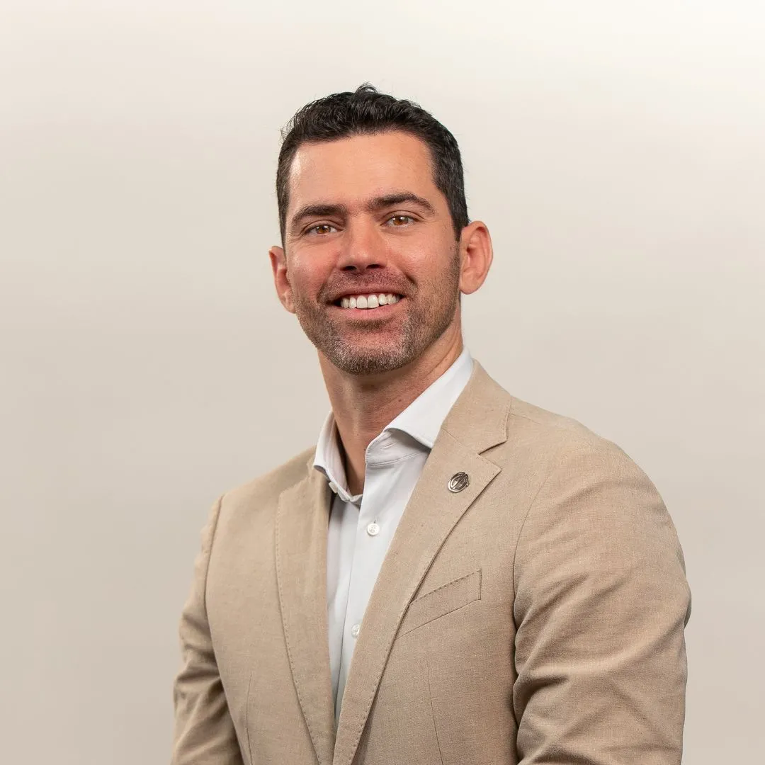 A man with short dark hair and a beard, wearing a beige suit jacket over a white shirt, smiles at the camera against a plain light background.