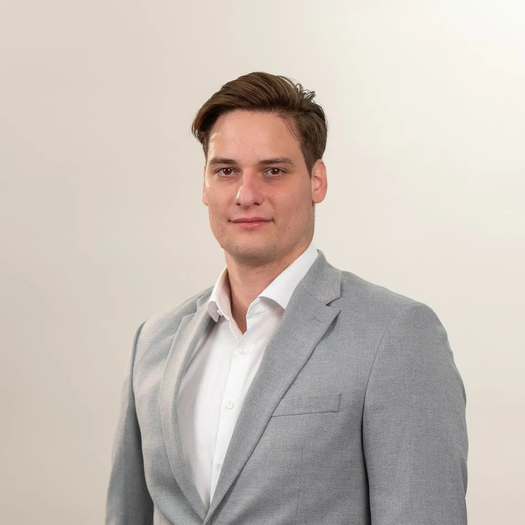 A man with short brown hair wears a light gray suit jacket over a white shirt, standing against a plain light background and looking at the camera with a neutral expression.