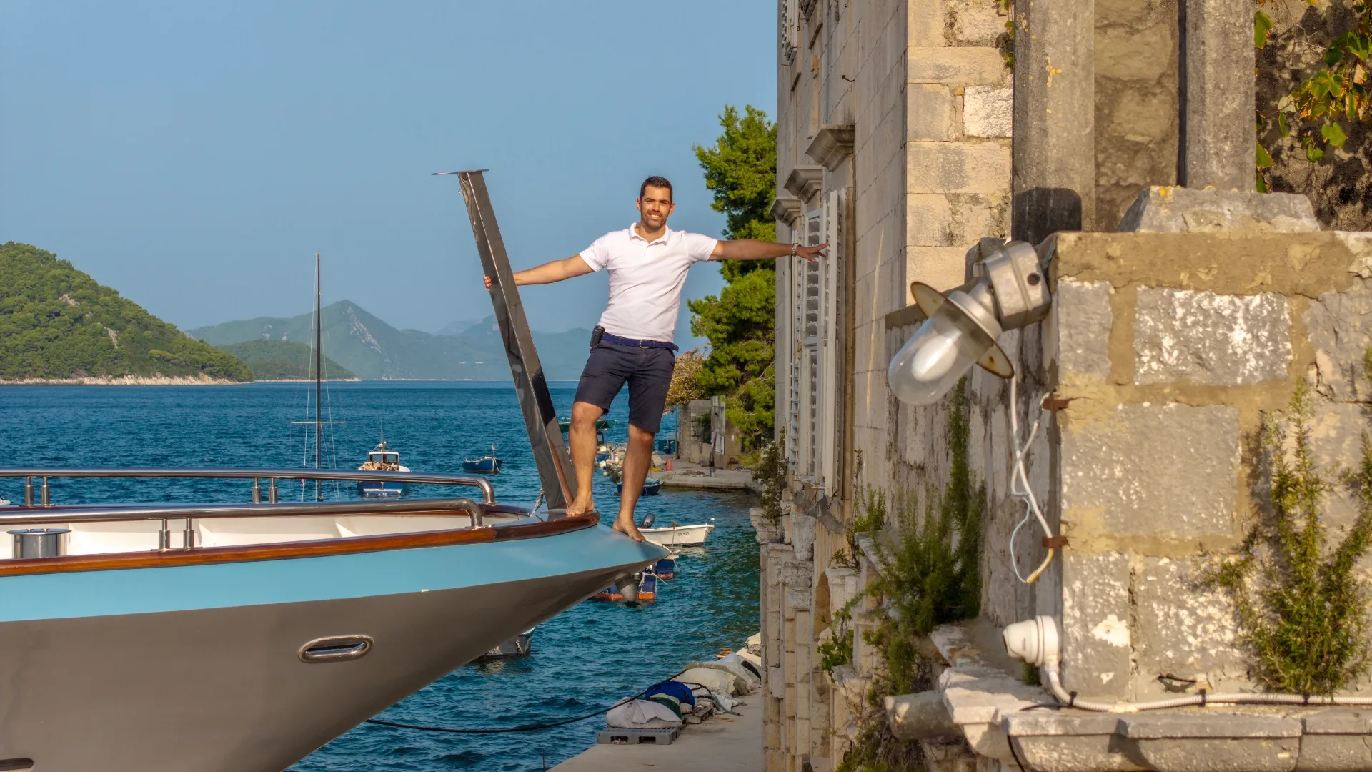 A man in a white shirt and navy shorts stands balancing on the bow of a yacht, arms outstretched, near a stone building by the sea with mountains and boats visible in the background.
