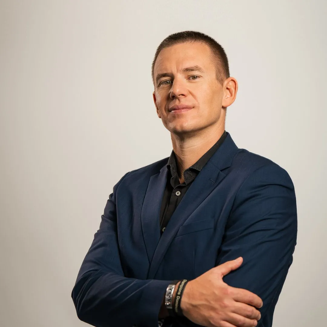 A confident man in a navy blue suit with arms crossed stands against a plain light background, looking directly at the camera with a slight smile.