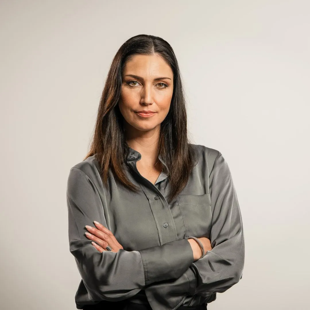 A woman with long dark hair wearing a silky gray button-up shirt stands with her arms crossed, looking confidently at the camera against a plain light background.