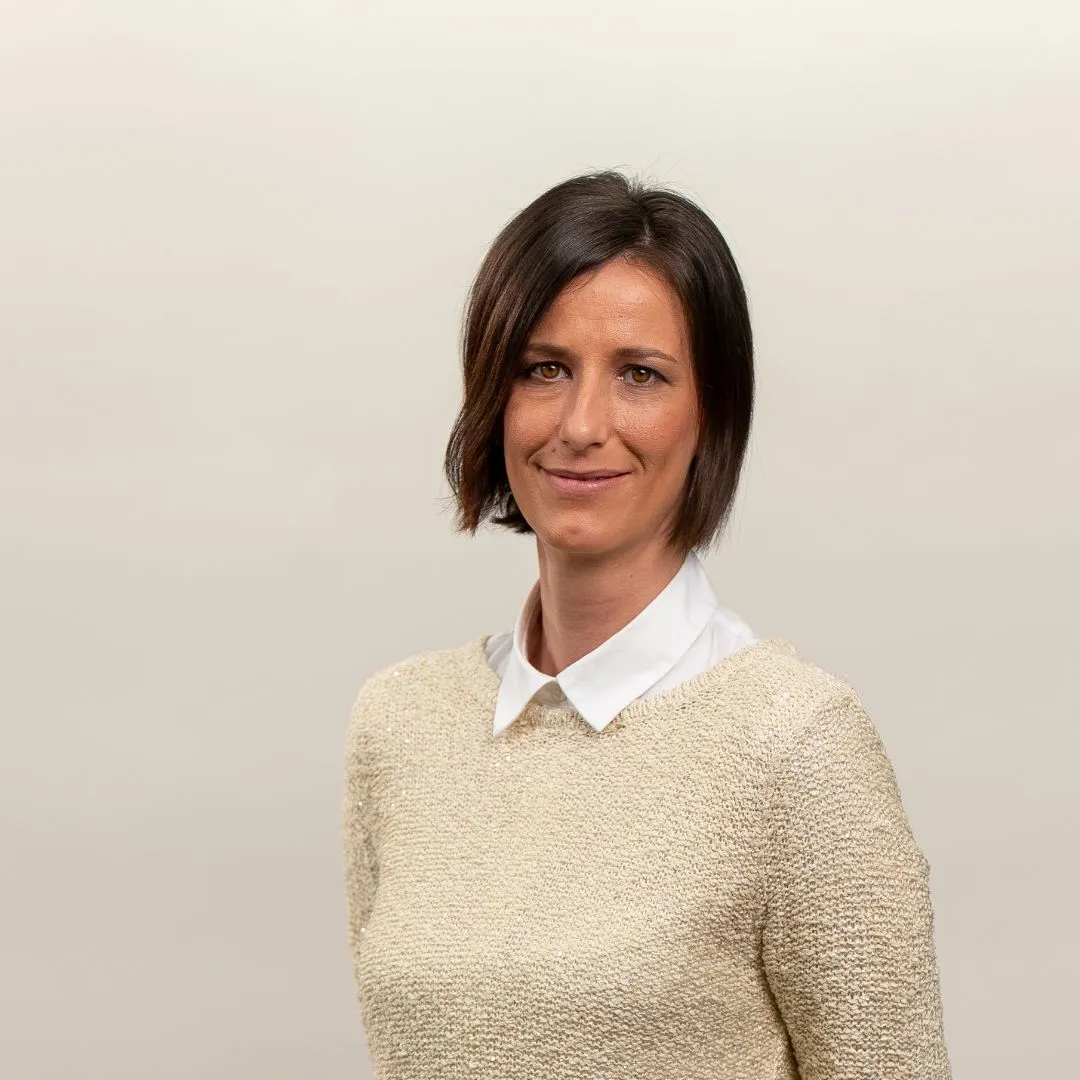 A woman with short dark hair, wearing a cream textured sweater over a white collared shirt, is smiling slightly while standing against a plain, light-colored background.