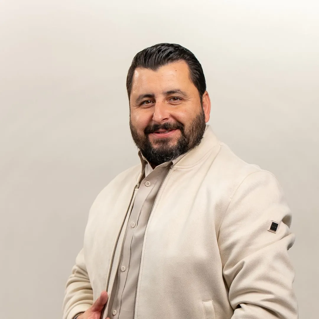 A man with dark hair and a beard smiles at the camera. He is wearing a light beige jacket over a matching shirt, standing against a plain, light-colored background.