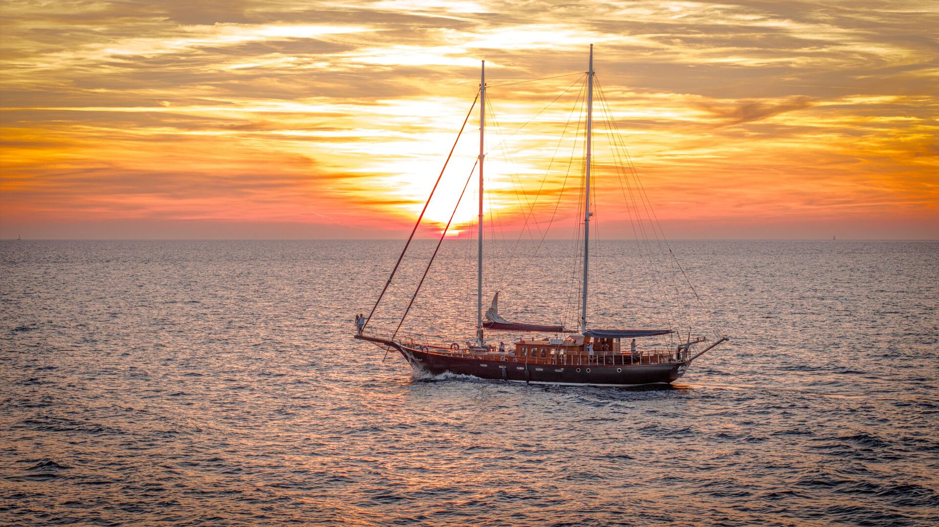 A sailboat glides on calm ocean waters at sunset, with vibrant orange and yellow clouds filling the sky and the sun low on the horizon.