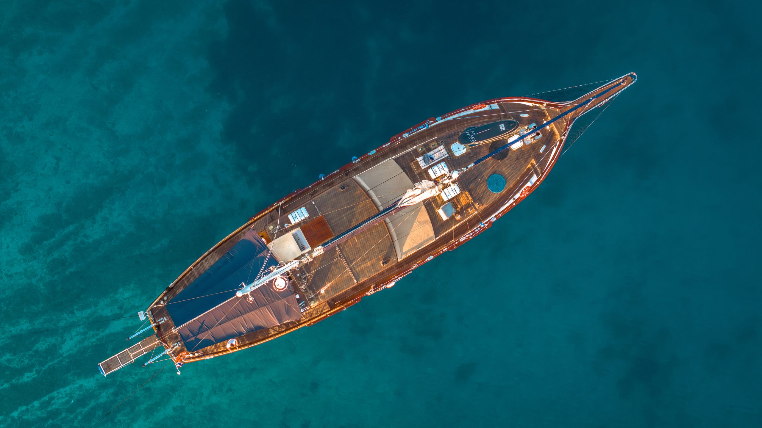 Aerial view of a wooden sailboat with white details floating on clear blue water, showing deck furniture and sails neatly arranged.