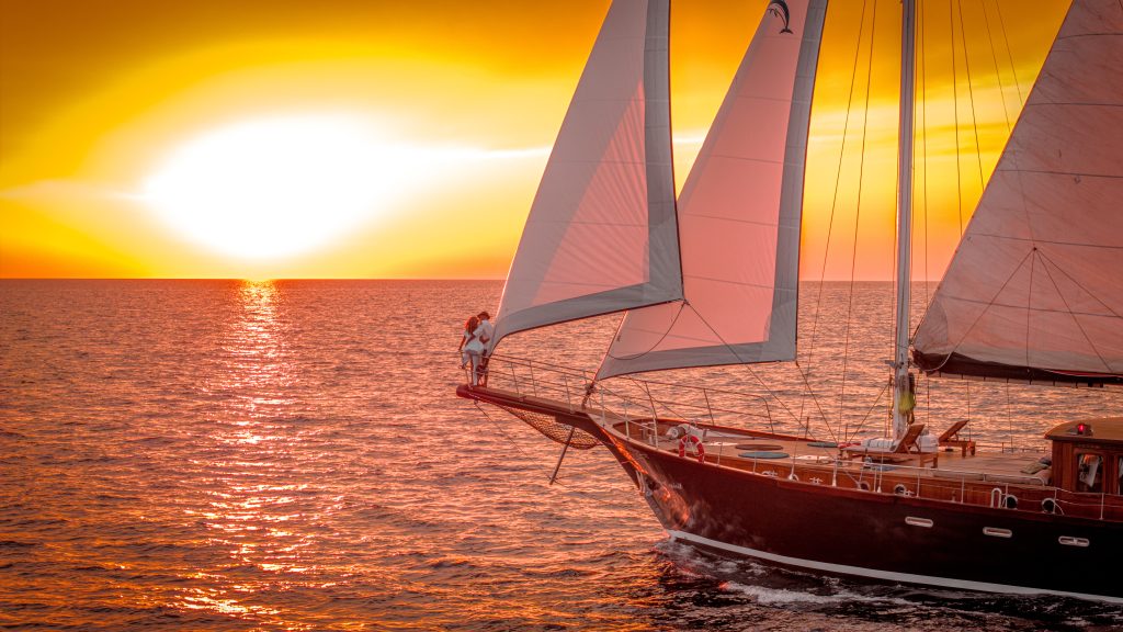 A sailboat glides on calm water at sunset, with two people standing at the bow, silhouetted against a vibrant orange and yellow sky.