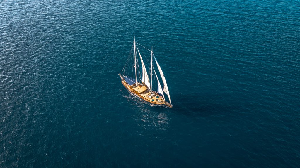 A wooden sailboat with white sails glides on calm, deep blue ocean water, viewed from above. The boat is surrounded by open sea with no land in sight.