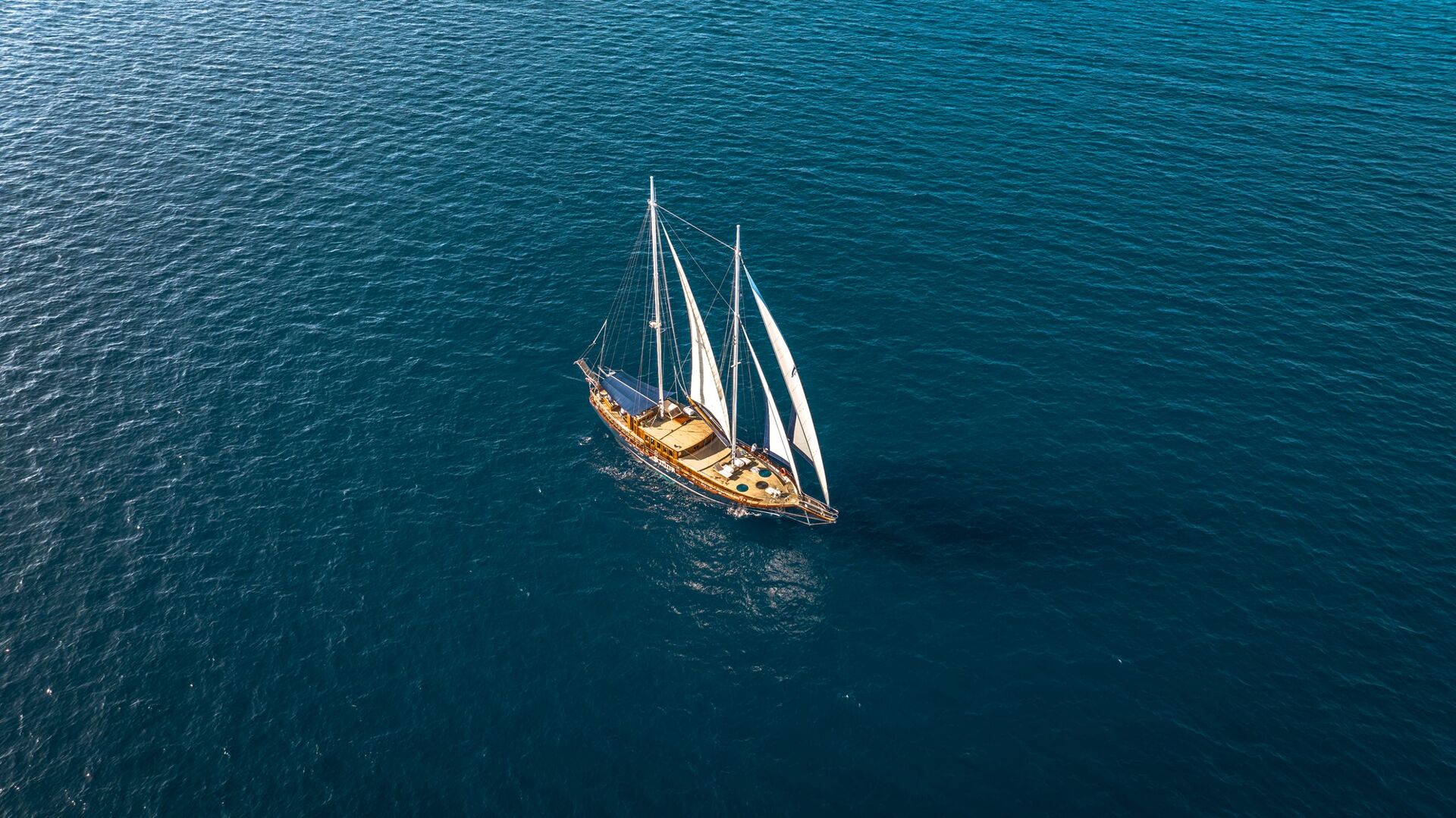 A wooden sailboat with white sails glides on calm, deep blue ocean water, viewed from above. The boat is surrounded by open sea with no land in sight.