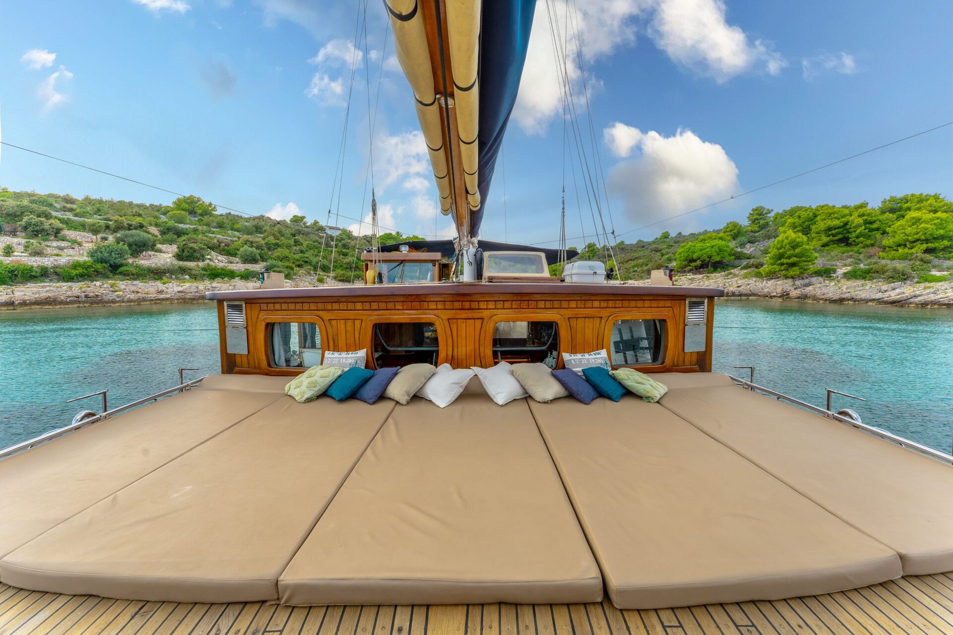 Sun loungers with colorful pillows line the deck of a wooden yacht, anchored in turquoise water near a lush, green, tree-lined shore under a partly cloudy sky.