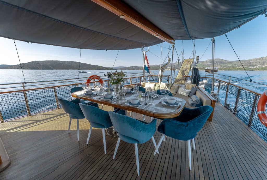 A yacht deck with a dining table set for eight, blue velvet chairs, cushioned bench, and flowers, under a canopy. The yacht is anchored on calm blue water, with hills and boats visible in the background.
