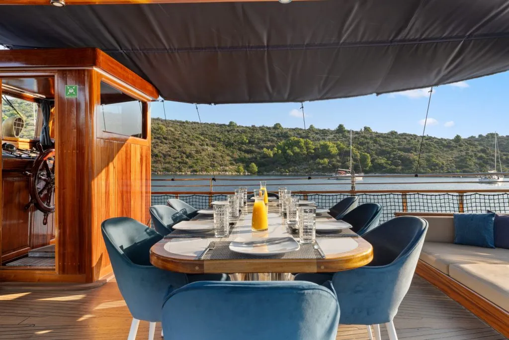 An outdoor dining table is set with plates, glasses, and condiments on a yacht. Blue chairs and cushioned benches surround the table, with a scenic view of water, hills, and anchored boats in the background.