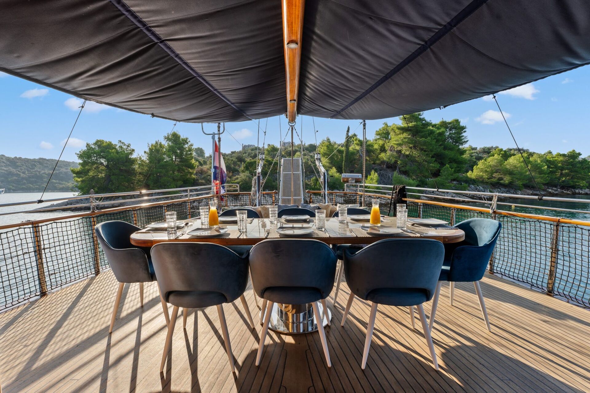 A dining table set for eight with blue chairs is arranged on the deck of a yacht, overlooking calm water and surrounded by lush green trees under a sunny sky.