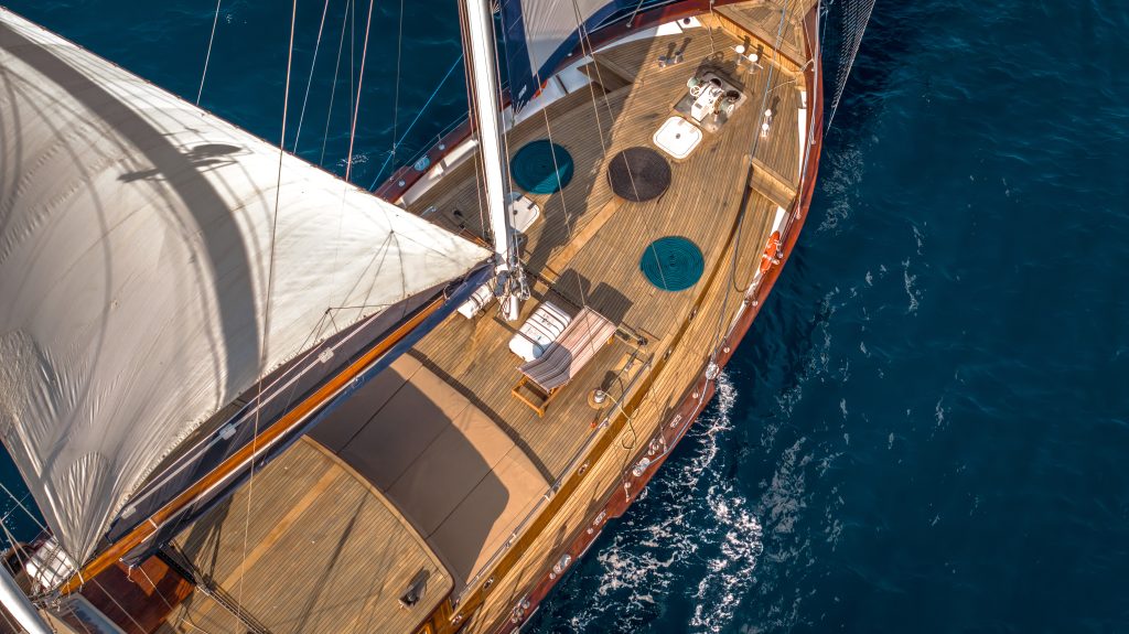 Aerial view of a wooden sailboat with white sails open, sailing on deep blue water. The deck features seating areas, sun loungers, and nautical equipment.