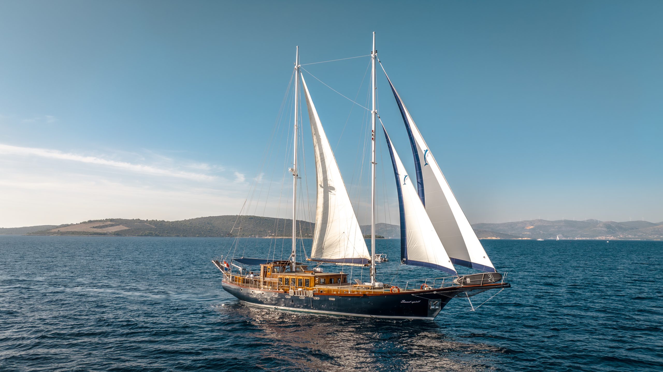 A large sailboat with two white sails glides across calm blue water under a clear sky, with a distant coastline and rolling hills visible in the background.