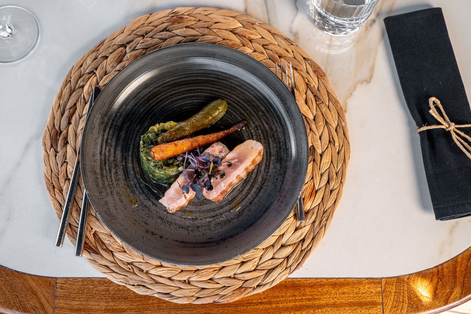 A dark plate on a woven placemat with slices of meat, mashed greens, a glazed carrot, a small cucumber, and microgreens. A glass of water and a black napkin tied with twine are beside the plate.