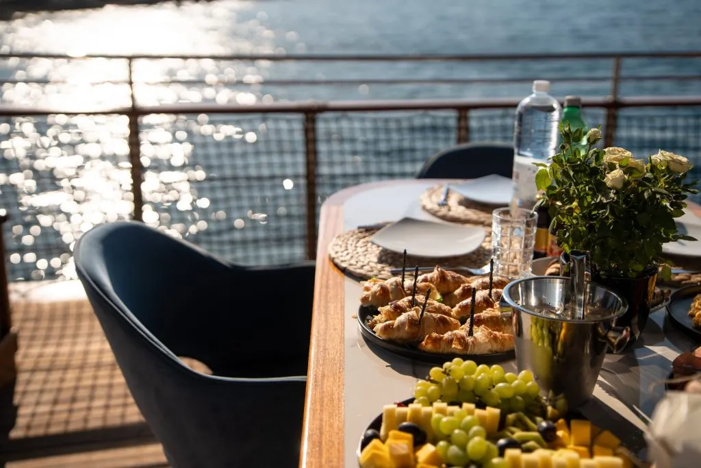 A sunlit table set with plates, pastries, fruit, water, and flowers overlooks sparkling water from a deck, with empty chairs inviting guests to enjoy the meal and scenic view.
