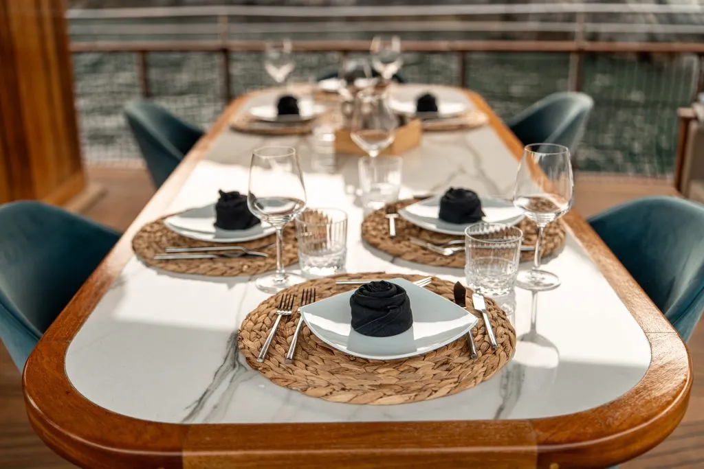 A close-up of an elegant outdoor dining table set with woven placemats, square white plates with black napkins, clear glasses, and cutlery, overlooking water with blurred background.