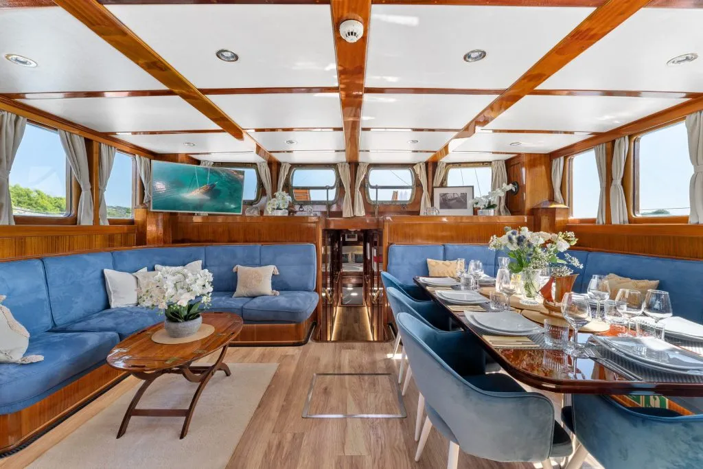 Luxurious yacht interior with polished wood finishes, blue upholstered seating, dining table set with elegant tableware, decorative flowers, large windows with curtains, and a TV mounted on the wall.